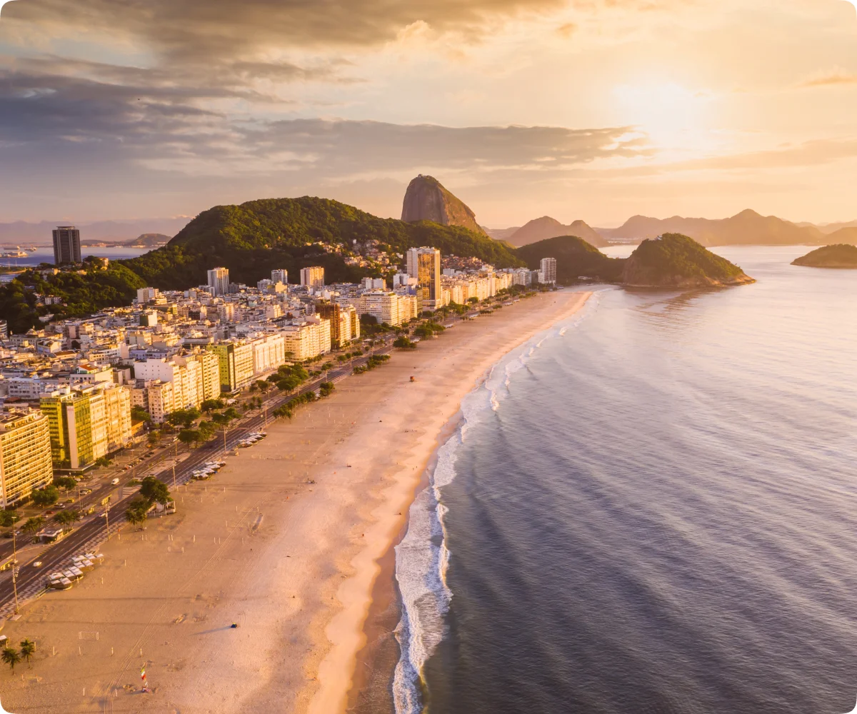 A imagem mostra uma bela vista aérea da cidade do Rio de Janeiro, destacando a famosa Praia de Copacabana ao amanhecer ou entardecer, com luz dourada iluminando a cena. A praia é ampla e extensa, com ondas suaves se quebrando na orla e poucas pessoas caminhando pela areia. À esquerda, vemos prédios altos alinhados, típicos da arquitetura urbana de Copacabana, enquanto ao fundo, o icônico Pão de Açúcar se eleva sobre a paisagem, reforçando o caráter natural e montanhoso da cidade. Pequenas ilhas e montanhas aparecem no horizonte, completando a vista deslumbrante do litoral carioca.